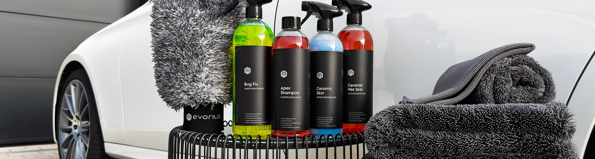 evorilo Fahrzeugpflegeprodukte vor weißem Fahrzeug – Bug Fix, Apex Shampoo, Ceramic Skin, Mikrofasertücher und Waschhandschuh im Detail