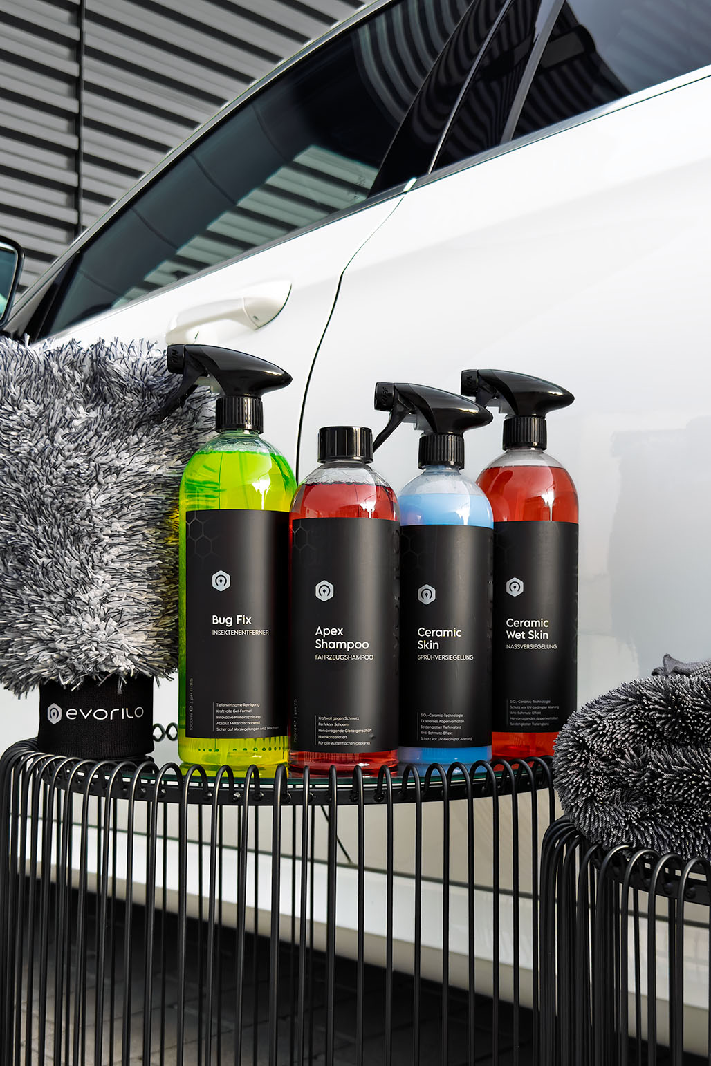 evorilo Fahrzeugpflegeprodukte vor weißem Fahrzeug – Bug Fix, Apex Shampoo, Ceramic Skin, Mikrofasertücher und Waschhandschuh im Detail